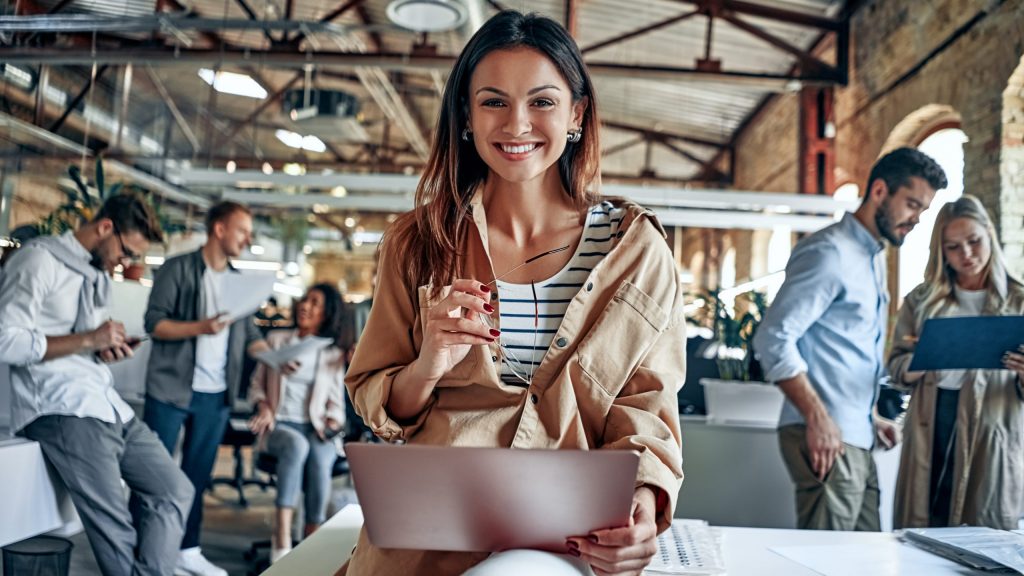 Girl with laptop smiling in an office with other people working in the background. 