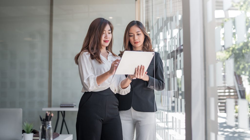 Two young Asian woman stand holding a tablet using SimVenture Validate by a window in their office.