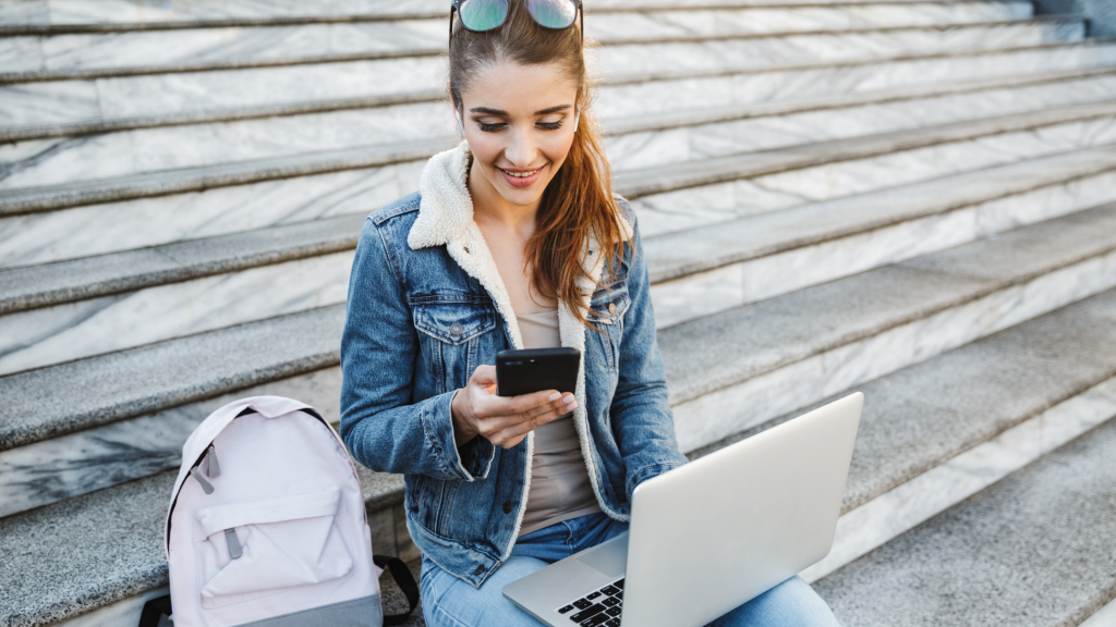 A young girl sitting on university steps looking at SimVenture Validate's interactive business model canvas on her phone and laptop.